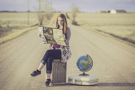 photograph of woman sitting on brown box beside desk globe on white briefcase center of road