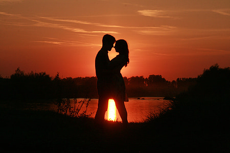silhouette of couple standing beside body of water against sun ray