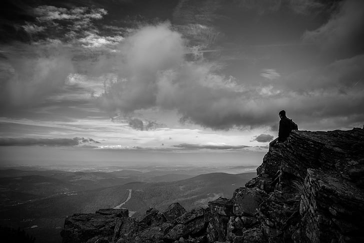 man sitting on rocky cliff looking at mountains in grayscale photography