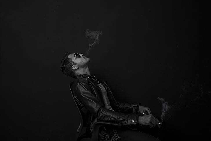 man wearing black leather jacket while blowing smoke on cigarette
