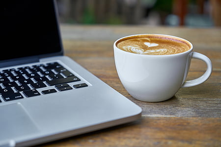 brown and white coffee in white ceramic mug beside of gray laptop computer