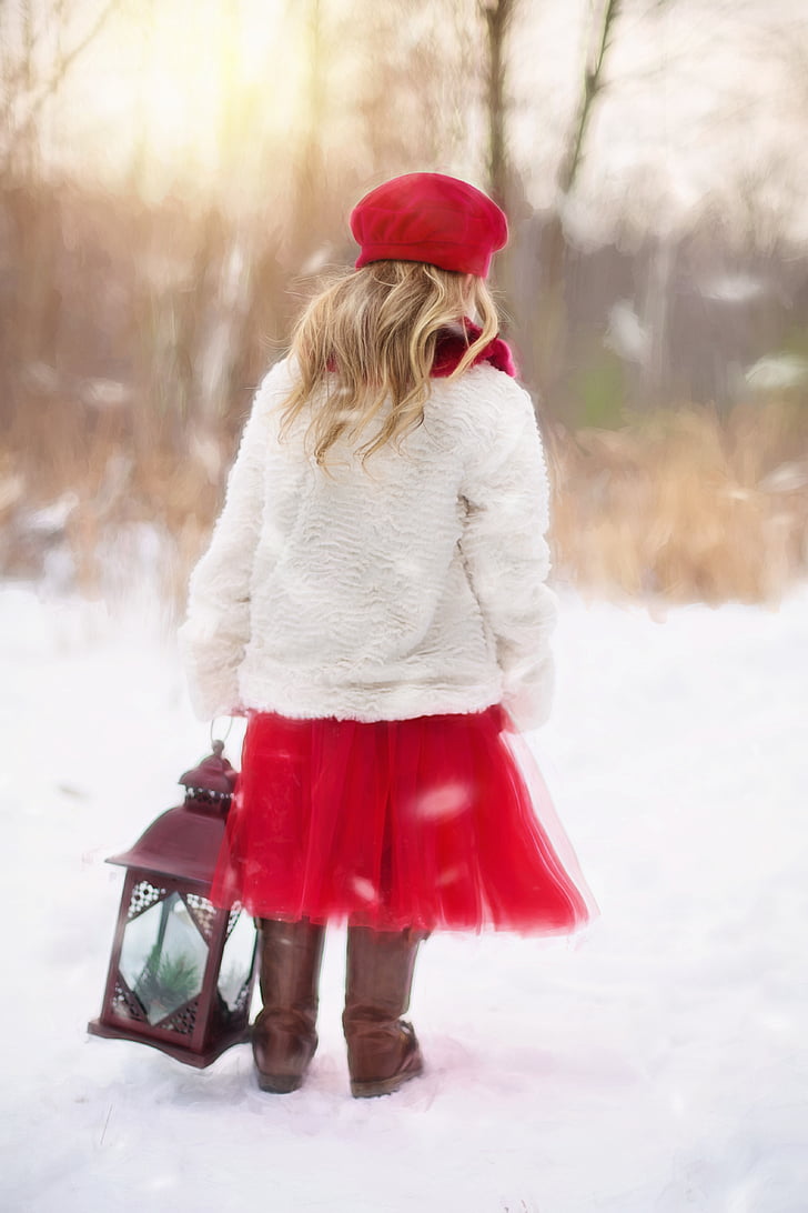 girl in white cardigan and red dress holding lantern standing on snow field during daytime